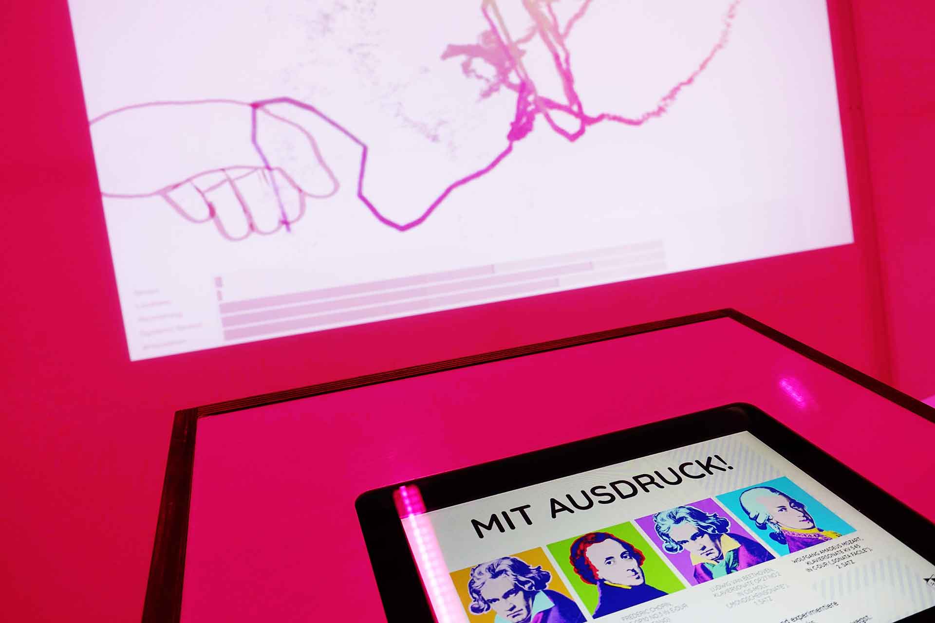 Das Touchscreen-Menü von Con Espressione zeigt die Songoptionen an. Im Hintergrund die Projektion einer Hand, die ausdrucksstarke Linien zeichnet.